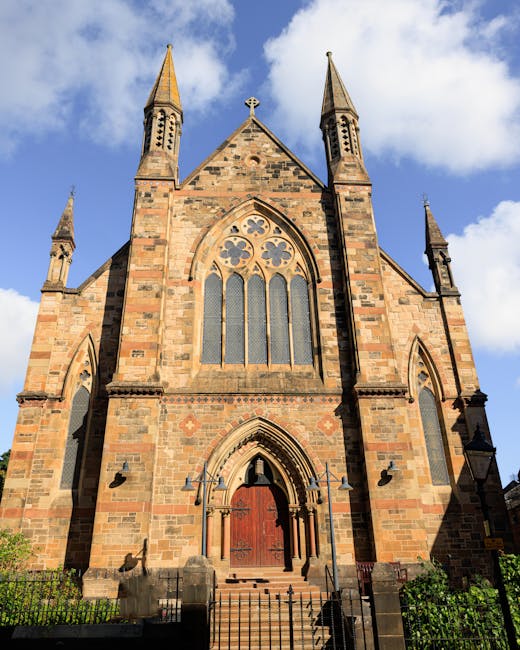 A tall, historic sandstone church on Church Street featuring Gothic architectural elements, including pointed arches and tall, narrow stained-glass windows. The building has four prominent spires and a large wooden door at the main entrance, accessible via a short set of stone steps with wrought iron railings. The facade is illuminated by natural sunlight, with a blue sky and some white clouds overhead, highlighting the textured stonework and intricate details. The surrounding area has small greenery and a black iron fence, emphasizing the church's prominent position on the street. The image reflects the church’s well-maintained, clean exterior, fitting into the context of community and heritage preservation, as highlighted in Paddington Carpet Cleaning’s surface cleaning and maintenance services for sacred and historic buildings.