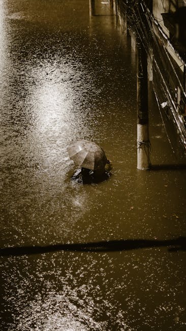 A flooded street at night with water covering the pavement and roadside; a person holding an umbrella is partially submerged in the floodwater near a building with dark exterior walls. The scene is illuminated by streetlights, creating reflections on the wet surfaces. Although this image is outside the typical interior cleaning context, it depicts the aftermath of flooding requiring emergency water removal and surface cleaning, which Paddington Carpet Cleaning offers in their emergency flood and spill cleaning services in Paddington (W2).