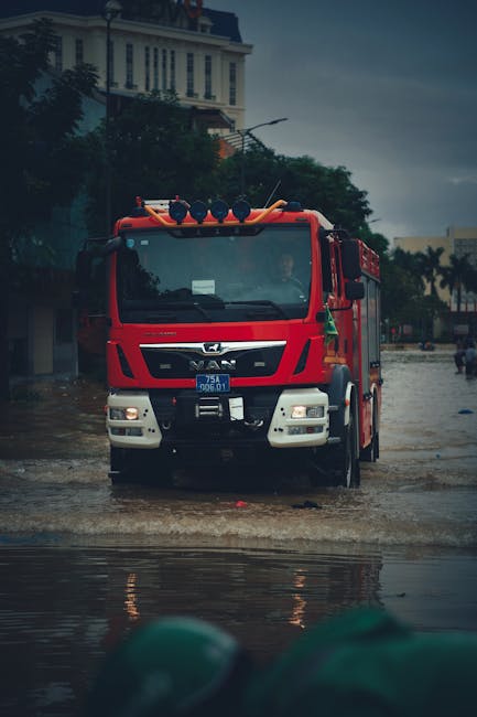 A red emergency flood response vehicle with blue flashing lights and equipment on top, situated on a flooded city street during heavy rain in Paddington. The street surface, made of asphalt, is submerged in water, reflecting the cloudy, dark sky. In the background, there are historic white buildings with decorative facades and trees, indicating an urban area affected by flooding. The scene captures the urgency of flood management and rescue efforts, demonstrating the importance of professional emergency cleaning and water removal services provided by Paddington Carpet Cleaning for property maintenance and hygiene during such incidents.