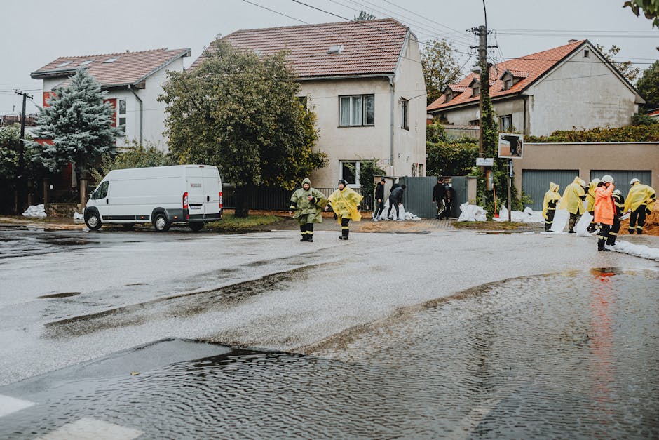 A flooded street at night with water covering the pavement and roadside; a person holding an umbrella is partially submerged in the floodwater near a building with dark exterior walls. The scene is illuminated by streetlights, creating reflections on the wet surfaces. Although this image is outside the typical interior cleaning context, it depicts the aftermath of flooding requiring emergency water removal and surface cleaning, which Paddington Carpet Cleaning offers in their emergency flood and spill cleaning services in Paddington (W2).