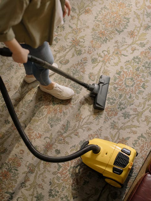A woman is engaged in deep surface cleaning of a traditional patterned area rug using a yellow canister vacuum cleaner with a flexible hose. She is standing on a wooden floor adjacent to a red leather