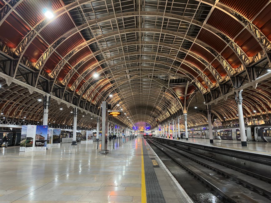 Interior view of Paddington Station with a modern high-speed train positioned on the platform. The station features an arched glass and metal roof allowing natural light to illuminate the polished stone floor. On the platform, passengers with luggage are waiting, some seated and others walking, while the station’s historic clock hangs above the platform. Paddington Carpet Cleaning's professional staff are not visible in this image, but the setting highlights the importance of maintaining clean, hygienic environments in transport hubs through expert cleaning and sanitisation services. The platform surface appears clean, dry, and well-maintained, emphasizing the importance of surface cleaning for hygiene and safety.